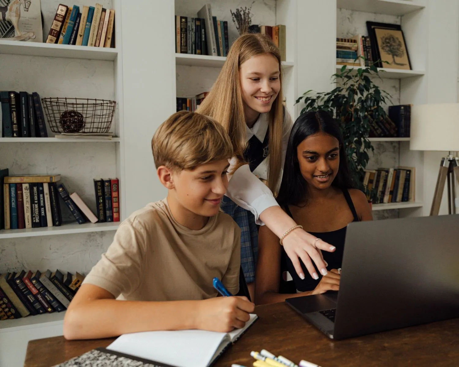 Three teenage learners studying together at a table, one writing notes while the others look on
