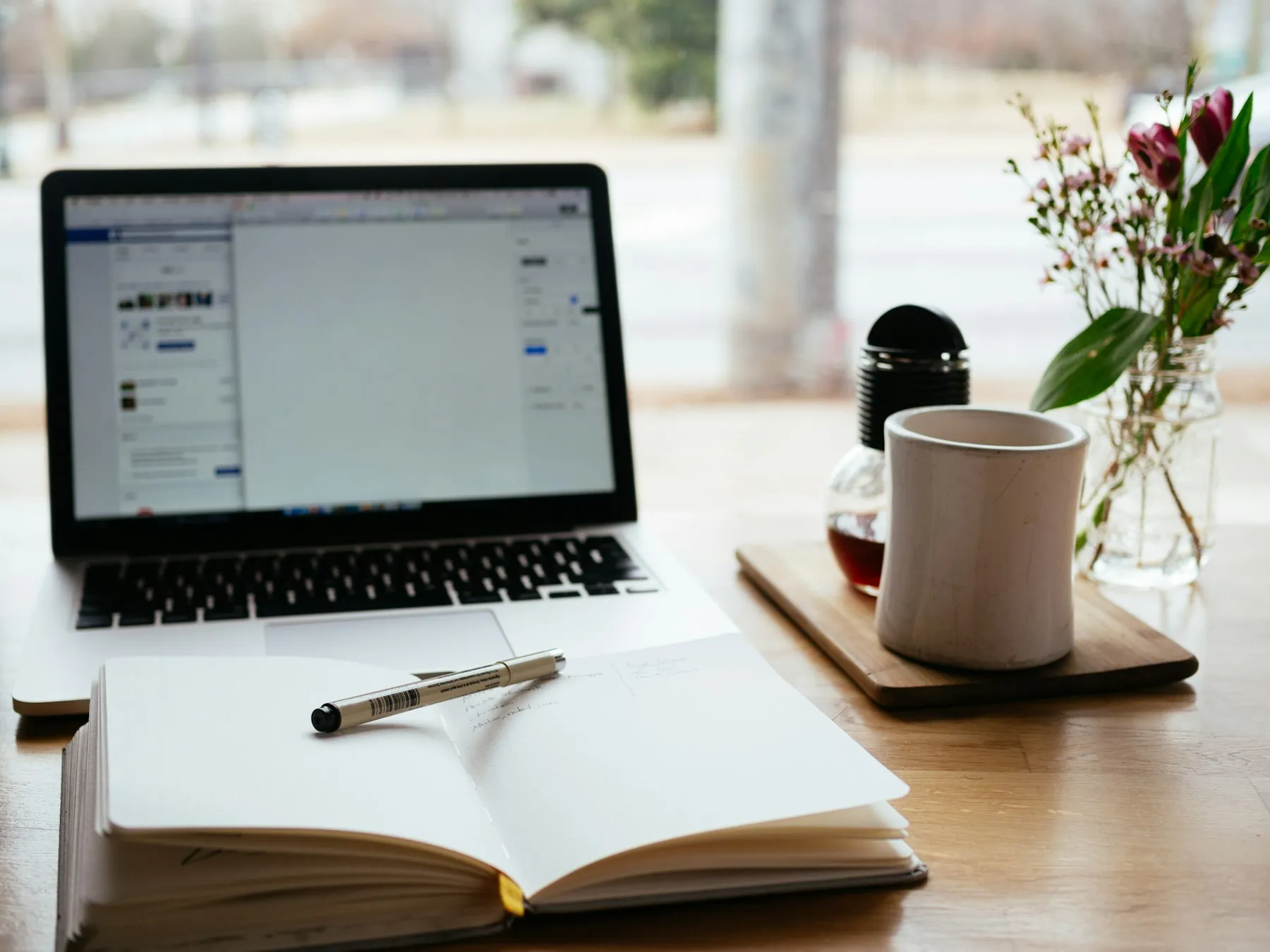 A laptop, an open notebook and a mug of coffee on a desk by a window
