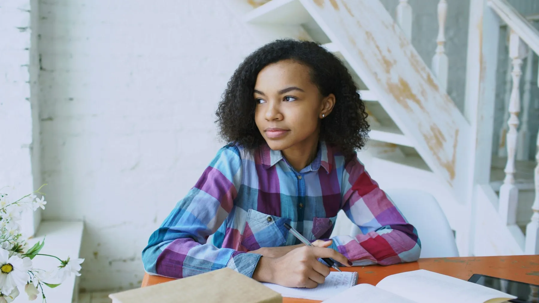 A teenage learner writing notes at a desk at home, looking up thoughtfully